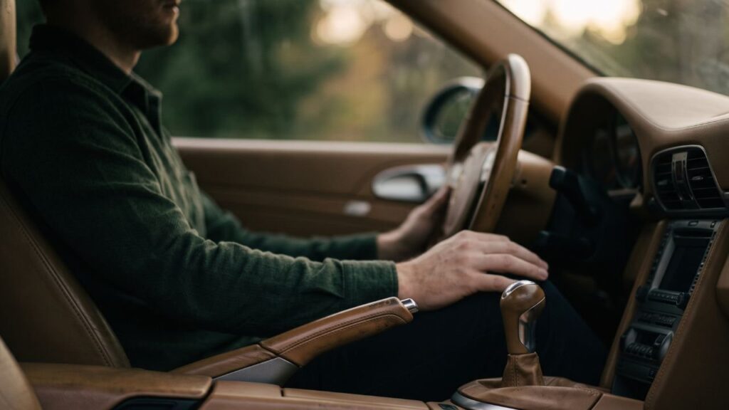 Mãos de um homem apoiadas no volante de um carro parado ao entardecer, freio de mão puxado, rosto fora do enquadramento